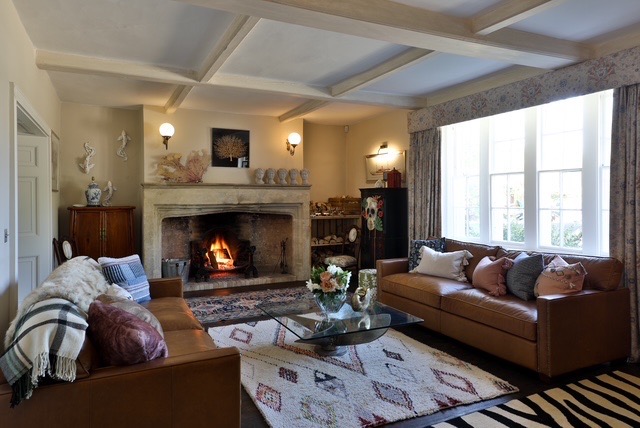 A cosy living room with leather sofas, patterned rugs, a glass coffee table with flowers, and a fireplace burning wood. Large windows let in natural light, and the room is decorated with vases and wall art. David Pearson Travel