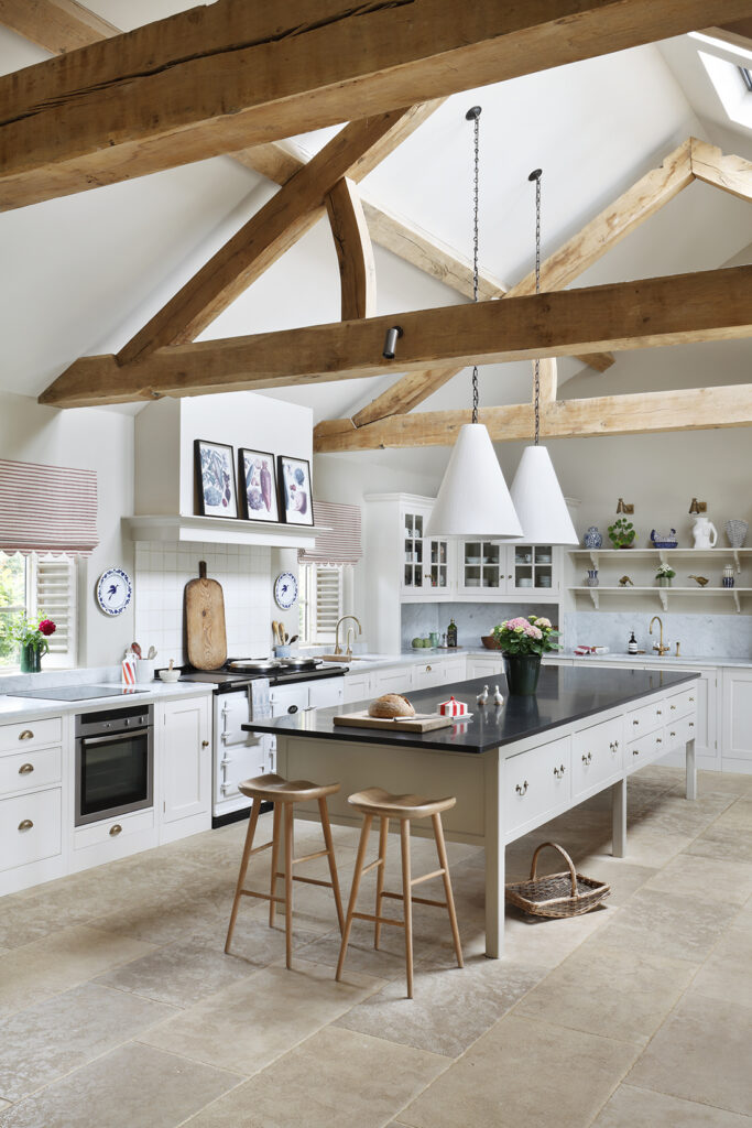 Spacious farmhouse kitchen with exposed wooden beams, white cupboards, a large black-topped island with stools, pendant lights, and open shelving decorated with crockery and plants. Natural light streams in from windows. David Pearson Travel