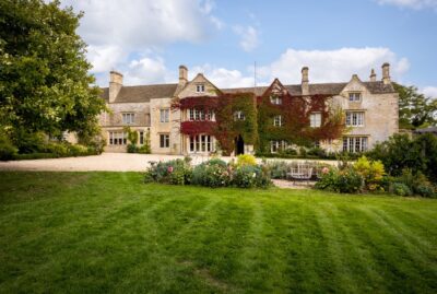 Large stone manor house with ivy growing on its central walls, tall chimneys, many windows, surrounded by landscaped gardens and a green lawn under a partly cloudy sky. David Pearson Travel
