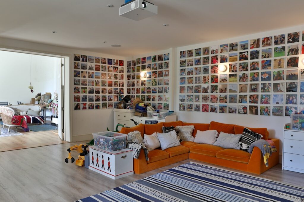 A cosy playroom with an orange corner sofa, striped rug, toy storage, and walls covered in colourful photo collages. An adjoining room with a bed and more toys is visible through open double doors. David Pearson Travel