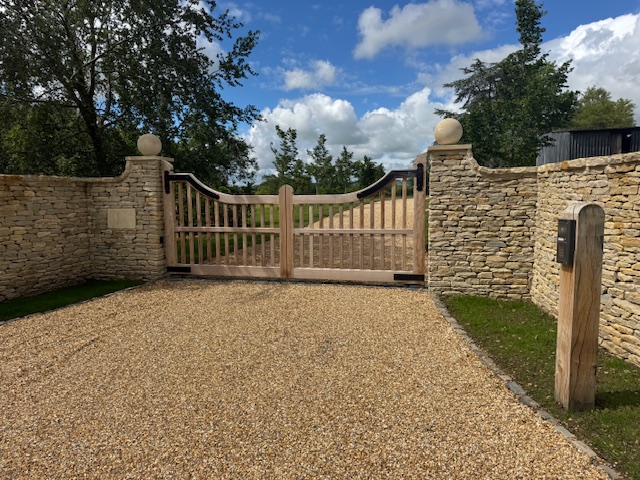A wide, wooden gate with stone pillars and walls stands at the entrance of a gravel drive, surrounded by green grass and trees under a partly cloudy sky. David Pearson Travel