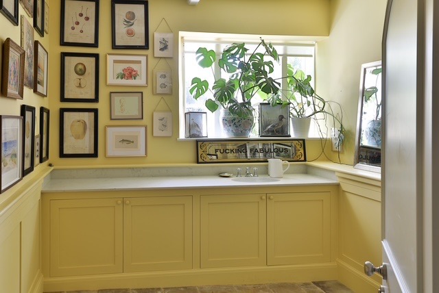 A bright yellow room with framed art on the walls, a worktop with a sink, a large plant by the window, and a decorative sign reading “Fucking Fabulous” displayed below the window. David Pearson Travel