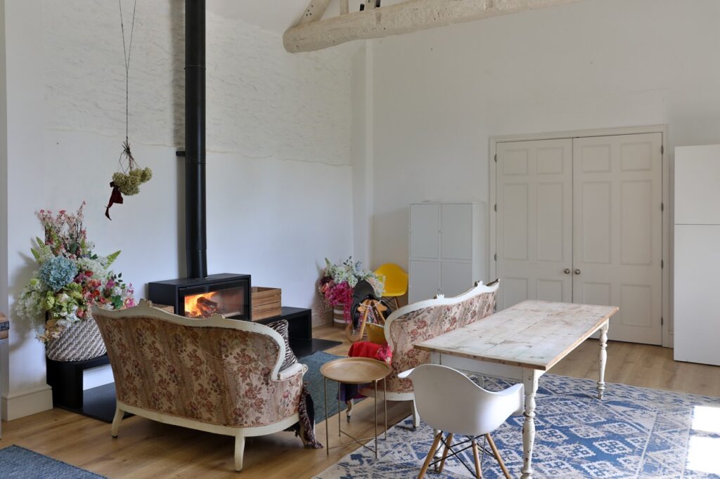 A cosy living room with floral sofas, a wood-burning stove, a wooden coffee table, a white Eames-style chair, floral arrangements, and a blue patterned rug on a wooden floor. White double doors are in the background. David Pearson Travel