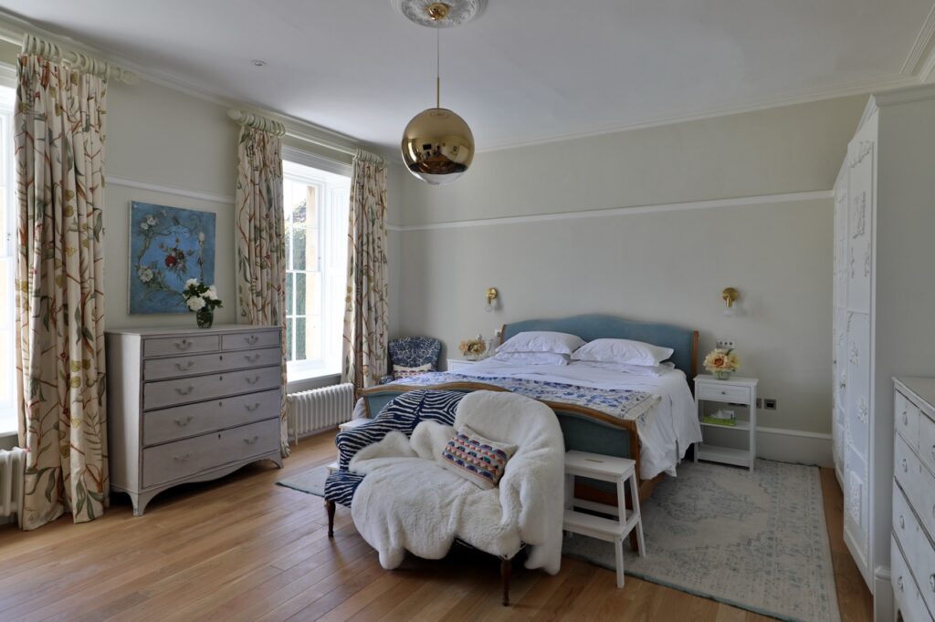 A bright bedroom with large windows, light curtains, a double bed with white bed linen, a gold pendant light, a white chest of drawers, a cosy armchair with throws, and wooden floors. A blue painting hangs on the wall. David Pearson Travel