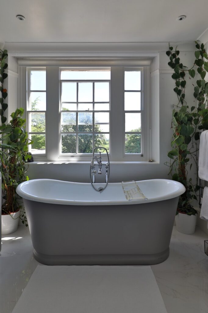A modern bathroom with a grey freestanding bath centred under a large window, surrounded by potted green plants and white walls, creating a bright and serene atmosphere. David Pearson Travel