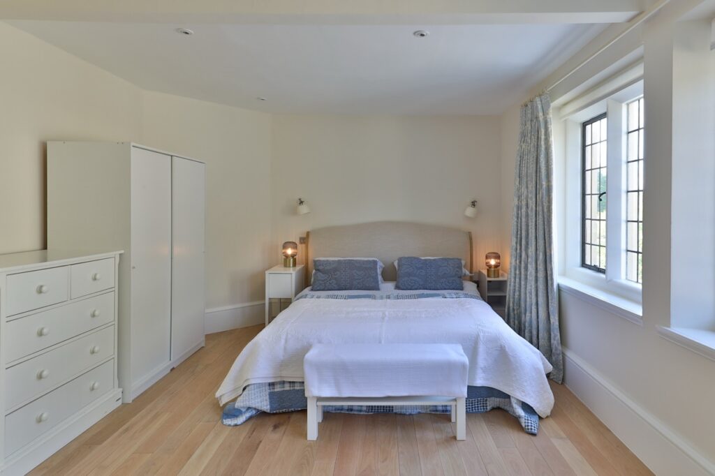 A bright, minimalist bedroom with a double bed, blue bedding, white furniture, wooden floor, two bedside tables with lamps, a large window with curtains, and a white wardrobe and chest of drawers. David Pearson Travel