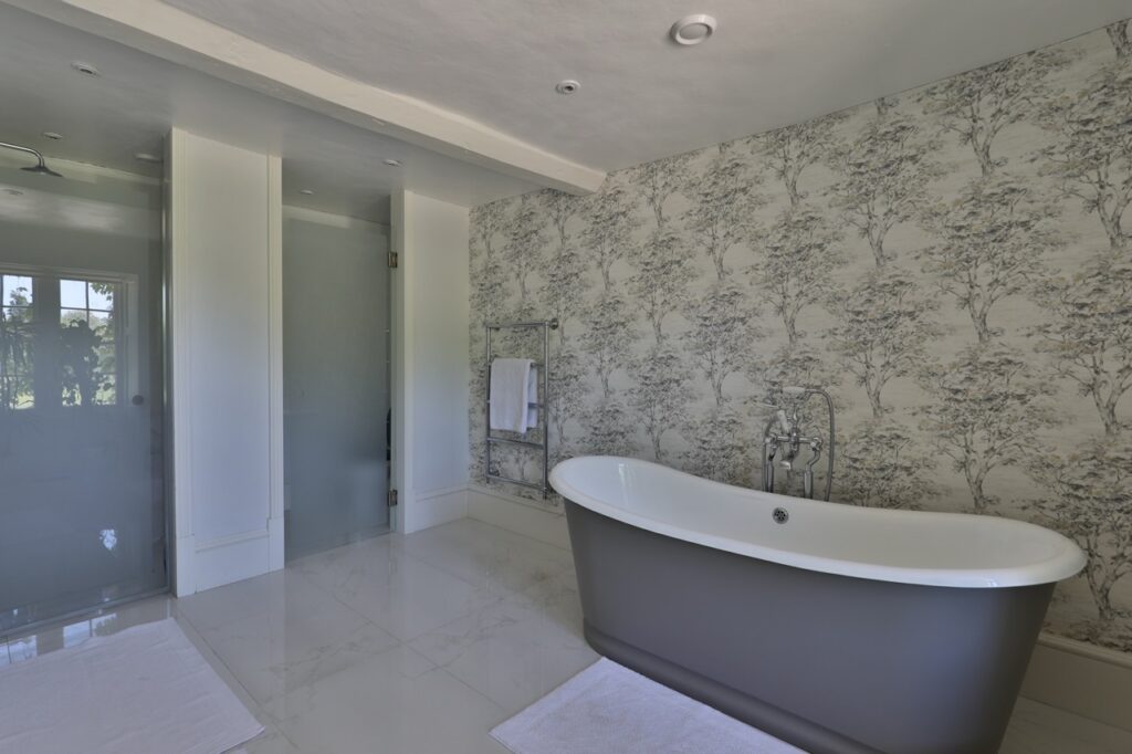 A modern bathroom with a freestanding grey bathtub, white tiled floor, patterned wallpaper, a towel rail with a white towel, and a glass shower enclosure. Natural light enters from a doorway in the background. David Pearson Travel