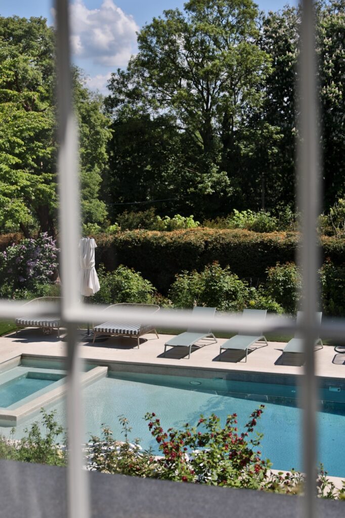 View through a window showing a poolside area with loungers and parasols, surrounded by lush greenery and tall trees under a partly cloudy sky. David Pearson Travel