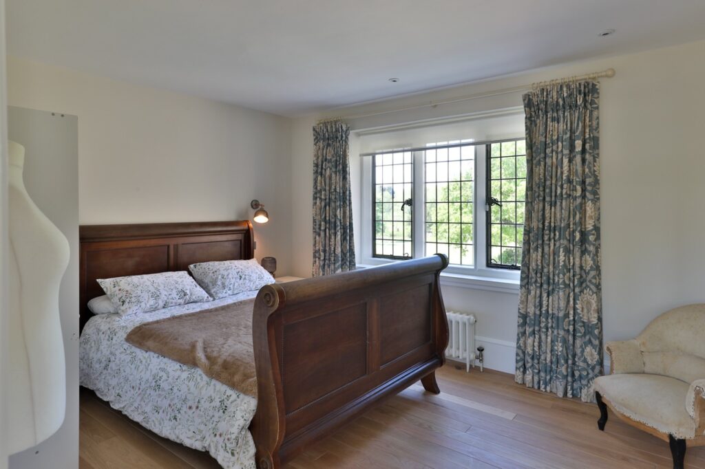 A bright bedroom with a wooden sleigh bed, floral bedding, a beige armchair, wooden floors, and a large window with patterned curtains letting in natural light. David Pearson Travel