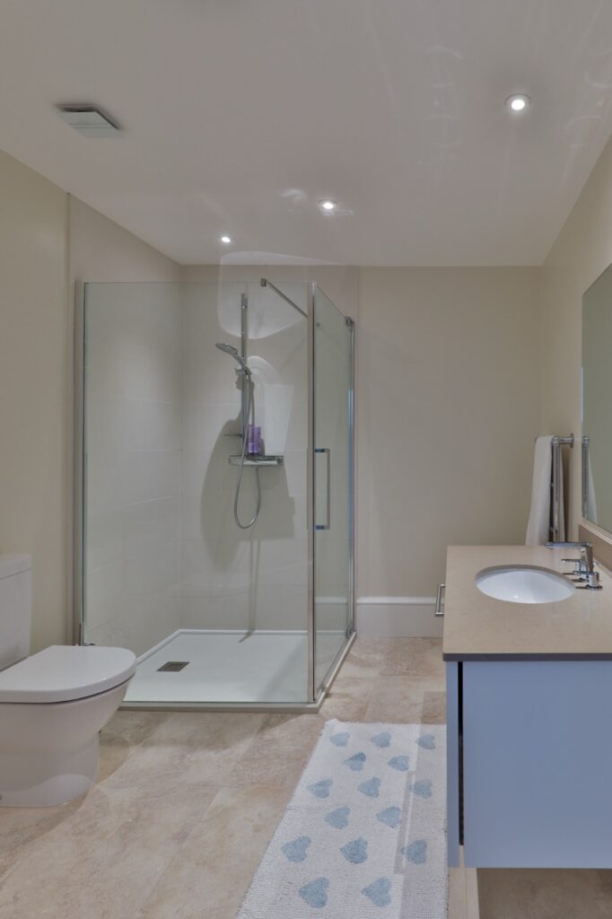 Modern bathroom with a glass shower enclosure, wall-mounted toilet, large vanity with basin, mirror, beige floor tiles, and a light-coloured rug with blue heart patterns. David Pearson Travel