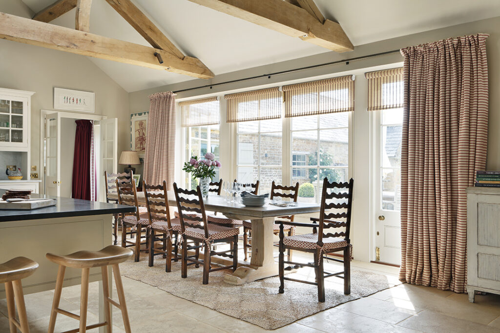 A bright dining room with exposed wooden beams, a long wooden table, eight chairs with patterned cushions, pink striped curtains, tall windows, and natural light filling the space. David Pearson Travel