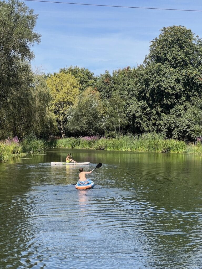 Two people kayak on a calm river surrounded by green trees and reeds under a clear blue sky. One kayaker is nearer the camera, paddling towards the other in the distance. David Pearson Travel