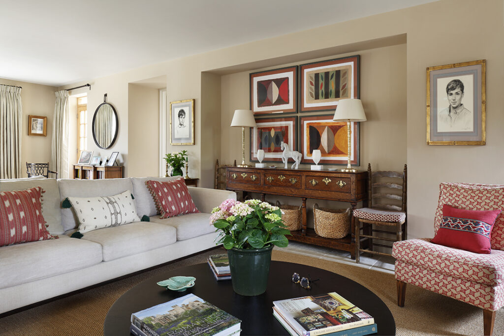A cosy living room with a beige sofa, patterned cushions, a round black coffee table with a potted plant and books, a red patterned armchair, and colourful modern art above a wooden sideboard. Framed portraits hang on beige walls. David Pearson Travel