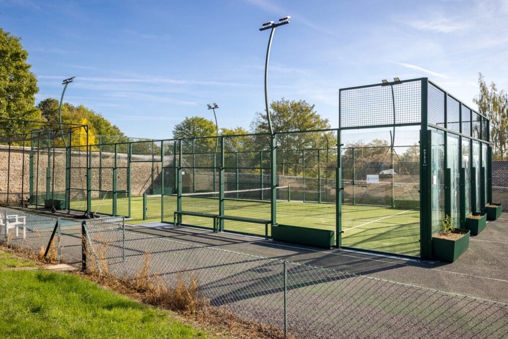 A fenced outdoor padel tennis court with artificial turf, surrounded by trees and blue sky, with empty seating and no players present. David Pearson Travel