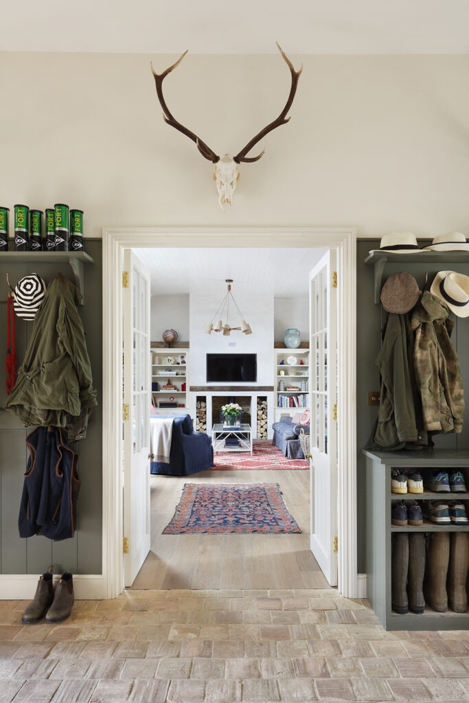 A well-organised mudroom with shelves for coats, boots, and hats leads to a bright living room. The living room has a TV, fireplace, bookcases, and a red patterned rug. Antlers are mounted above the doorway. David Pearson Travel
