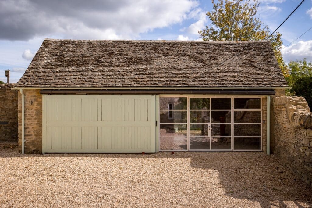 A stone garage with a shingle roof features a large pale green sliding wooden door on the left and a glass panelled door on the right, set on a gravel drive under a partly cloudy sky. David Pearson Travel
