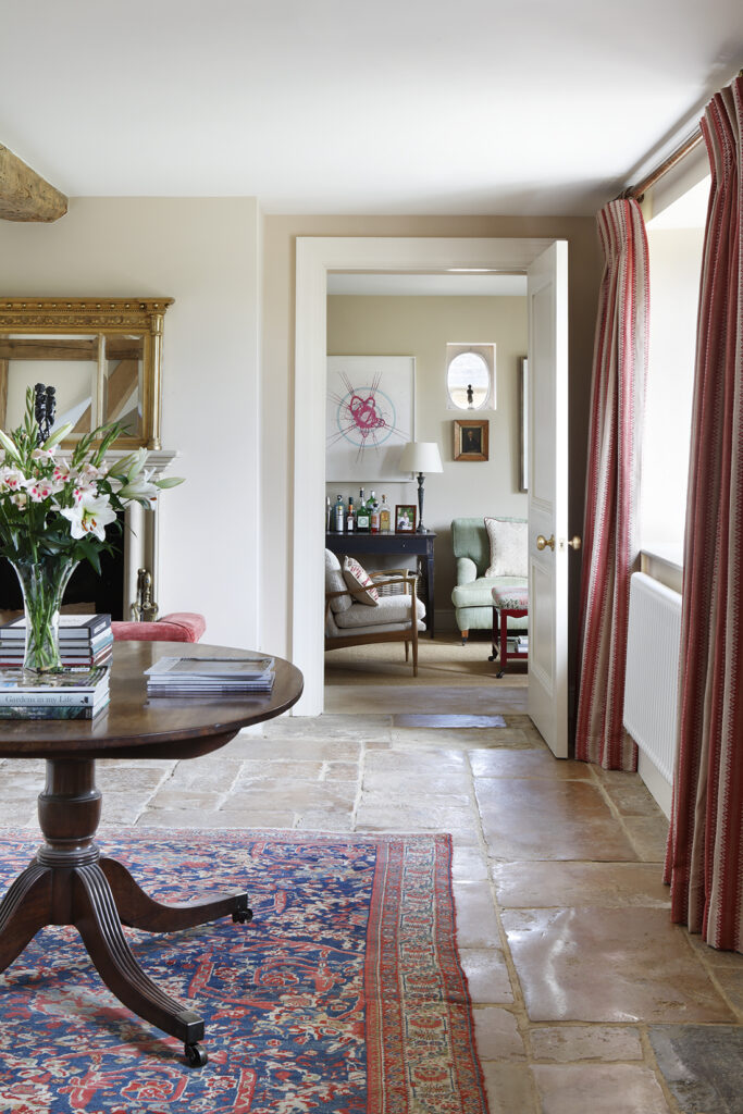 A cosy living area with stone tiled floors, a patterned rug, and a round wooden table topped with flowers and books. Through an open door, another room with armchairs, a drinks trolley, and wall art is visible. Red curtains frame a window. David Pearson Travel