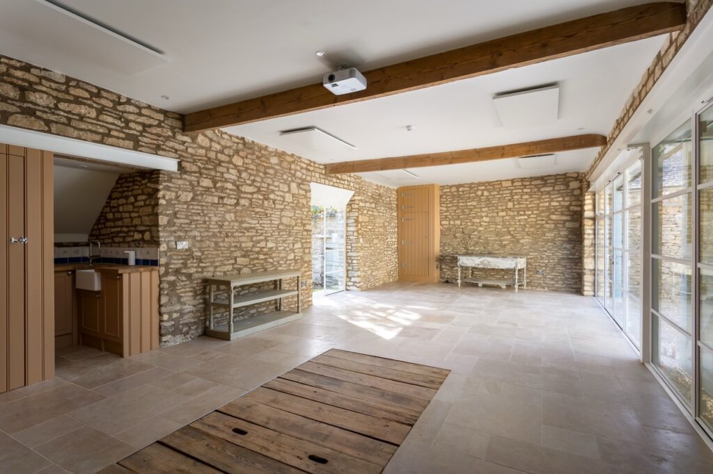A spacious room with stone walls, large windows, wooden beams, and a tiled floor. There is a wooden trapdoor on the floor, a bench, a table, and an open doorway leading to a small kitchen area. Natural light fills the space. David Pearson Travel