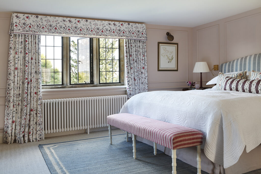 A cosy bedroom with pink walls, floral curtains, a large bed with white bedding and striped pillows, a pink upholstered bench, a framed picture, a rug, and a window letting in natural light. David Pearson Travel