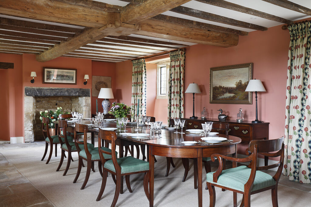 A traditional dining room with exposed wooden beams, a long polished wooden table laid for a meal, green-upholstered chairs, a stone fireplace, floral curtains, and artwork on coral-pink walls. David Pearson Travel