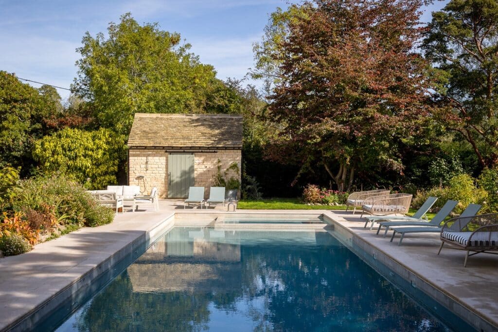 A serene outdoor swimming pool surrounded by loungers and greenery, with a small stone shed and large trees in the background under a clear sky. David Pearson Travel