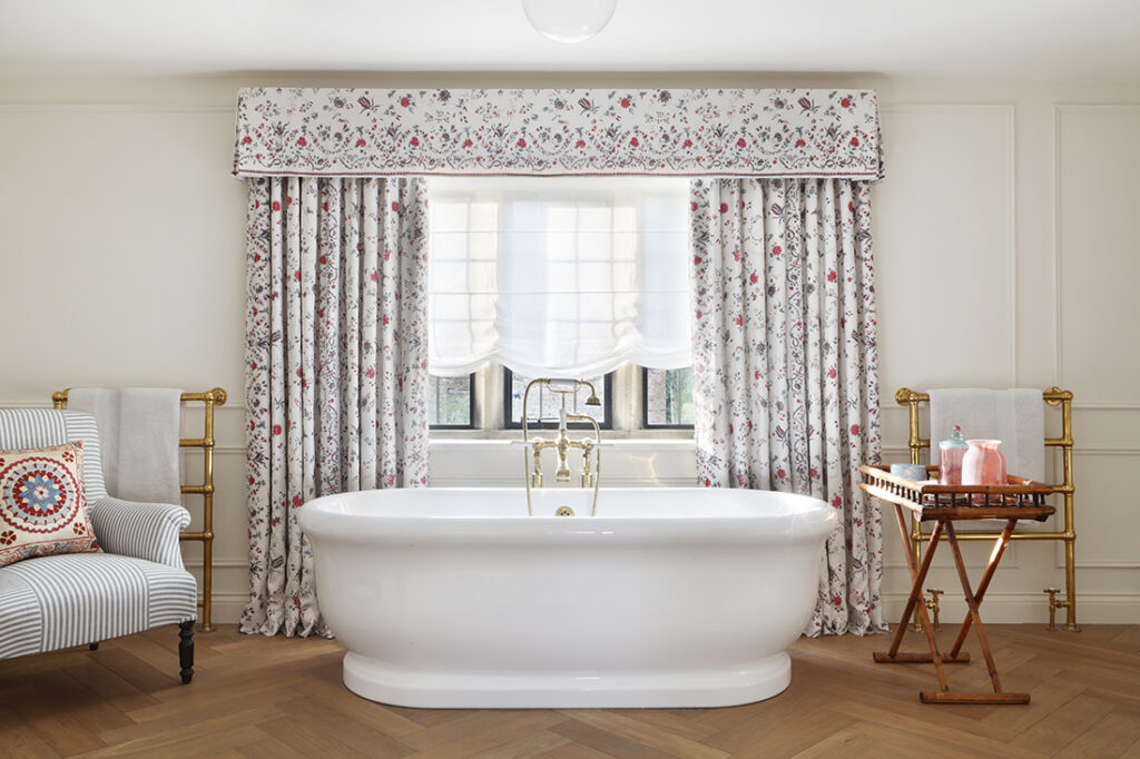 A white freestanding bath sits in front of a window with floral curtains. A wooden tray holds bottles and towels. There are gold towel rails and a striped chair with a colourful round cushion on the left. David Pearson Travel