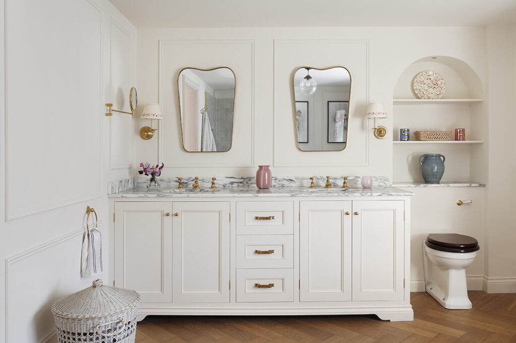 A bright, elegant bathroom with a double-sink vanity, gold fittings, two mirrors, marble worktop, decorative shelves with vases, a brown toilet, and a laundry basket on a herringbone wooden floor. David Pearson Travel