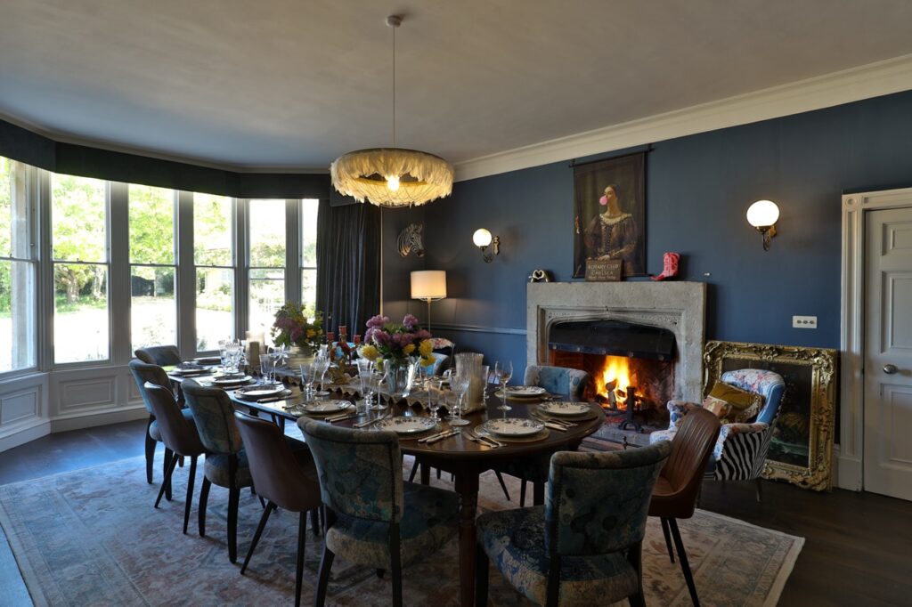 A cosy dining room with a long table set for a meal, blue walls, a fireplace with a fire burning, large bay windows, and a chandelier above. Flowers and glassware decorate the table, creating an inviting atmosphere. David Pearson Travel